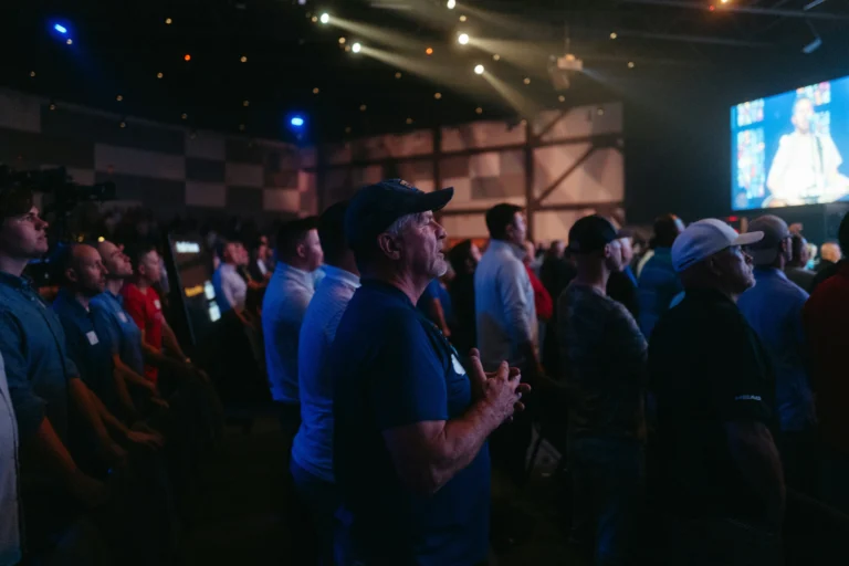a large worship center with people standing and singing. There is a screen in the background indicating a guitar player with a microphone is leading worship.