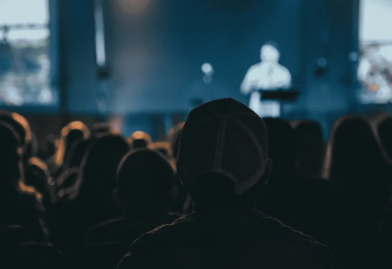 set in a modern church, the foreground shows the backs of many church goers, seated, and the background shows a blurred preacher