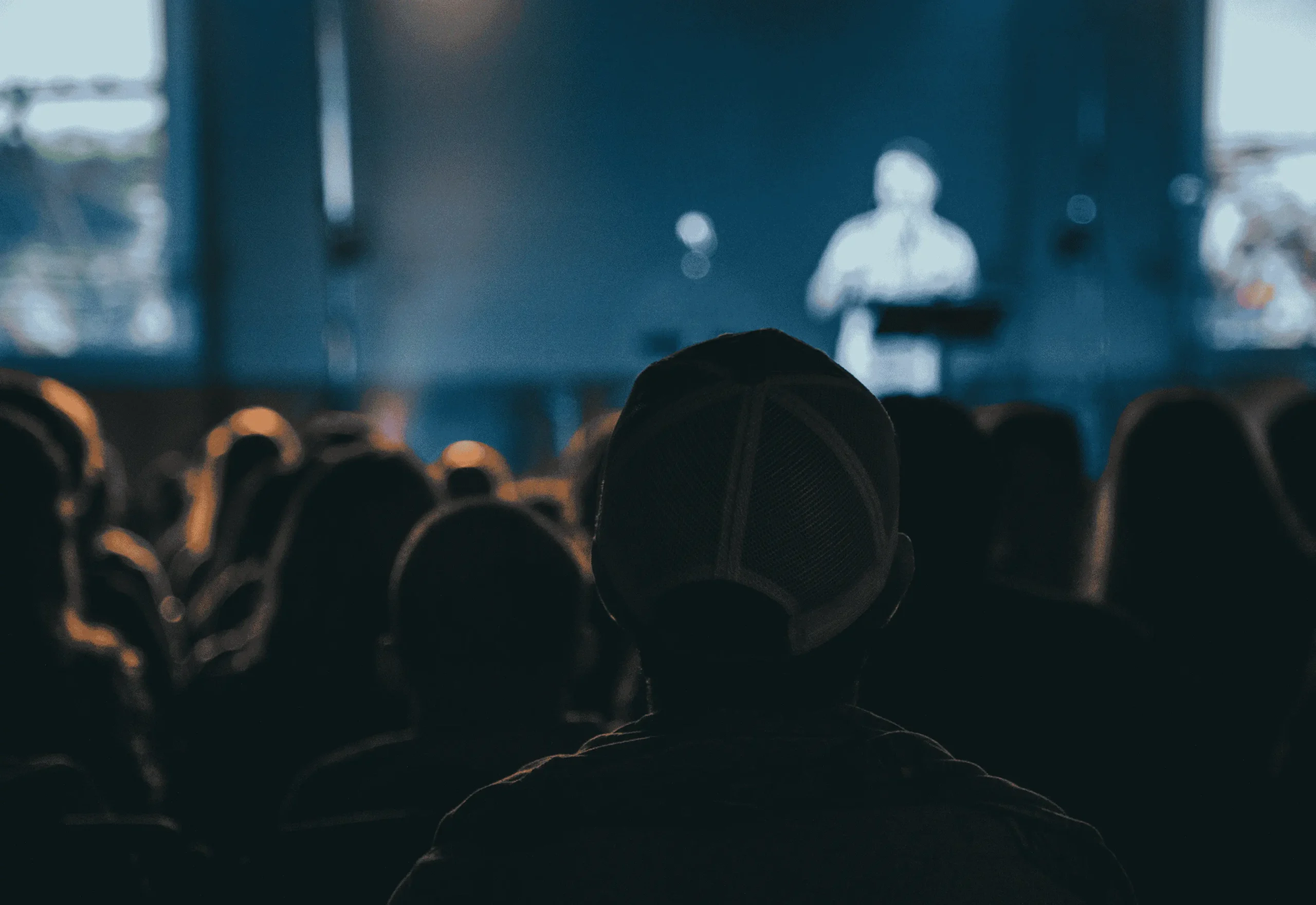 set in a modern church, the foreground shows the backs of many church goers, seated, and the background shows a blurred preacher